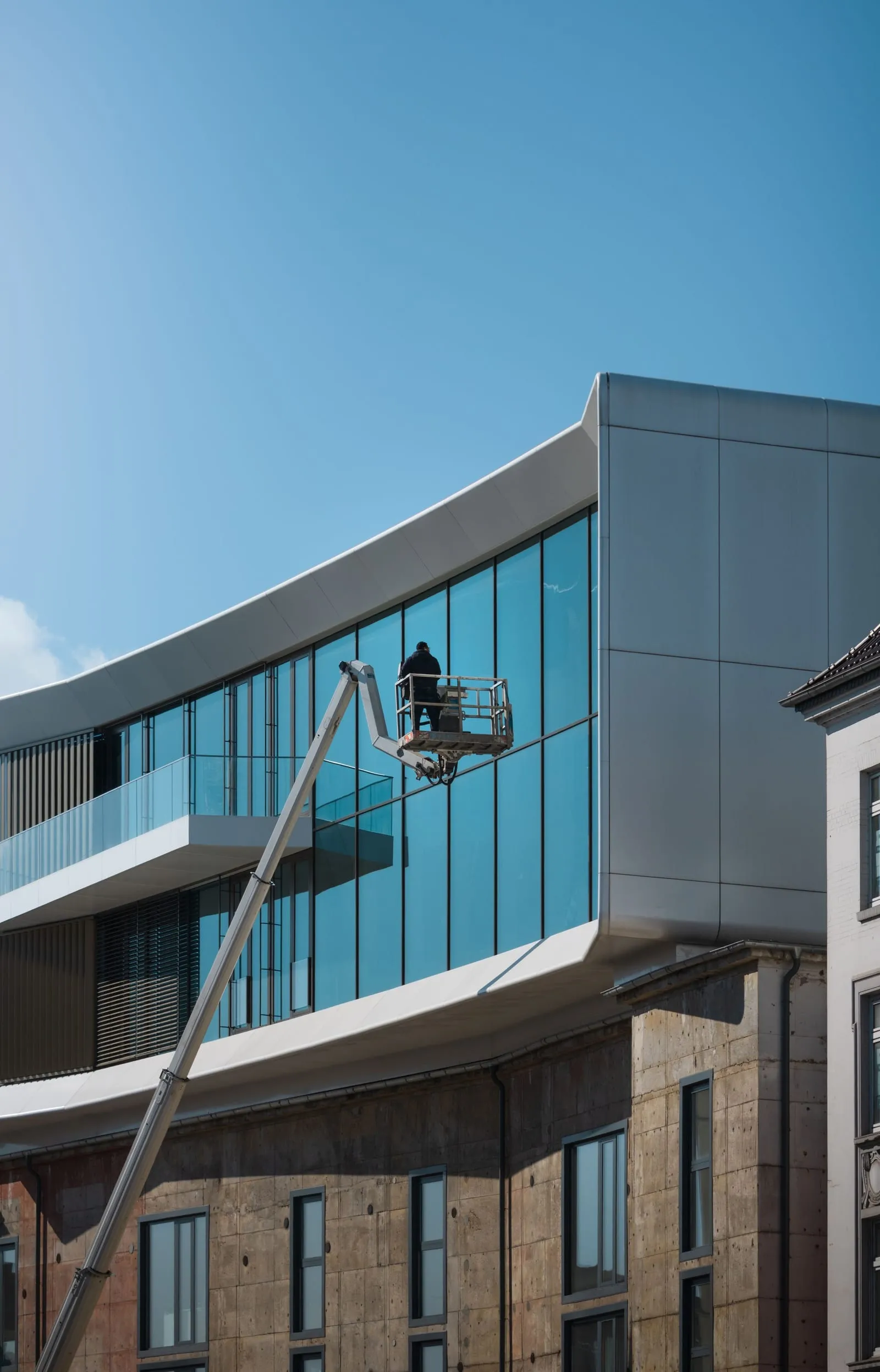 Fensterreiniger in einer Arbeitsbühne vor der gebogenen Glasfassade eines Bürohauses, blauer Himmel