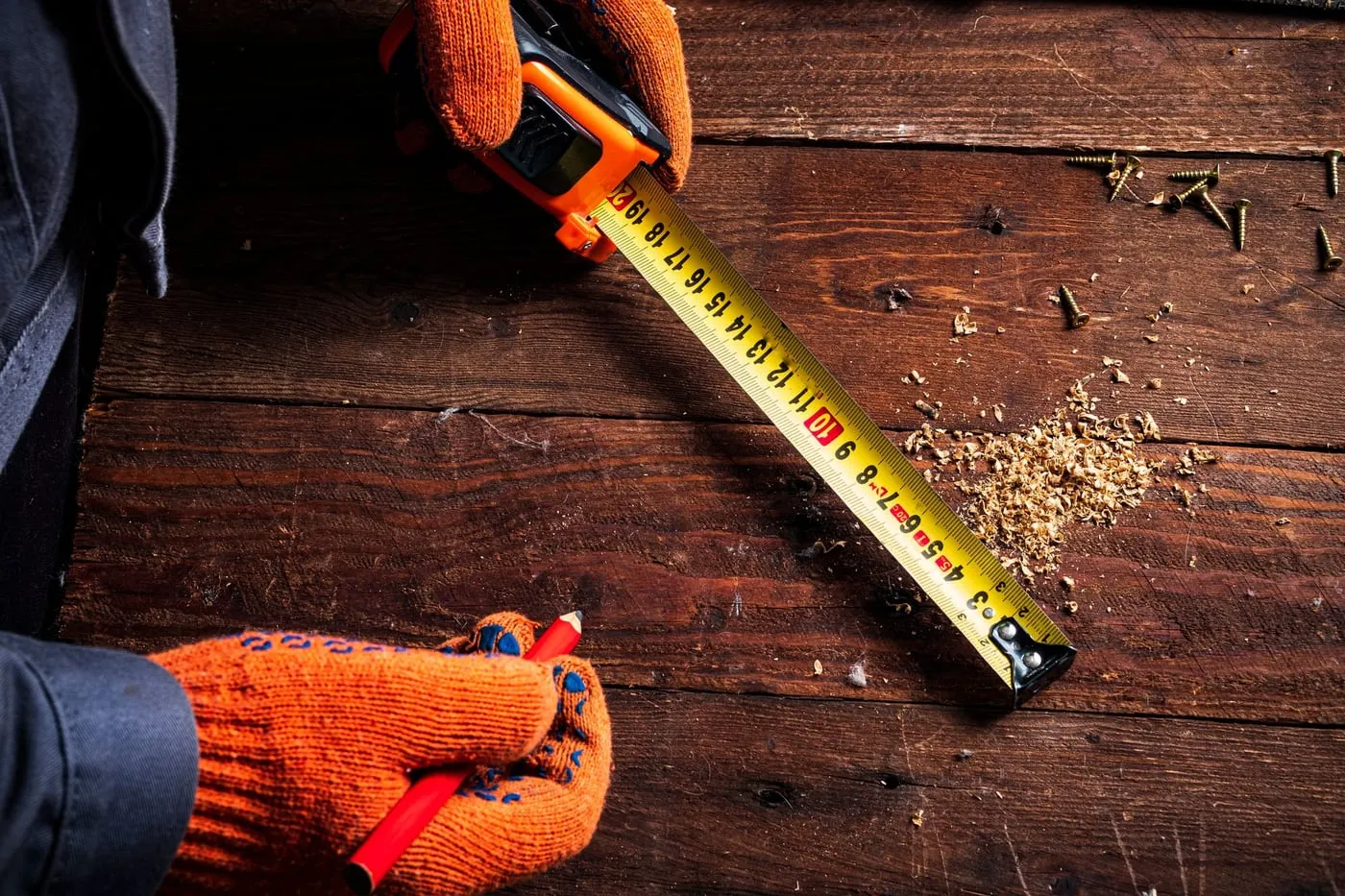 Hände in orangefarbenen Arbeitshandschuhen messen mit Maßband und Bleistift auf Holzdielen