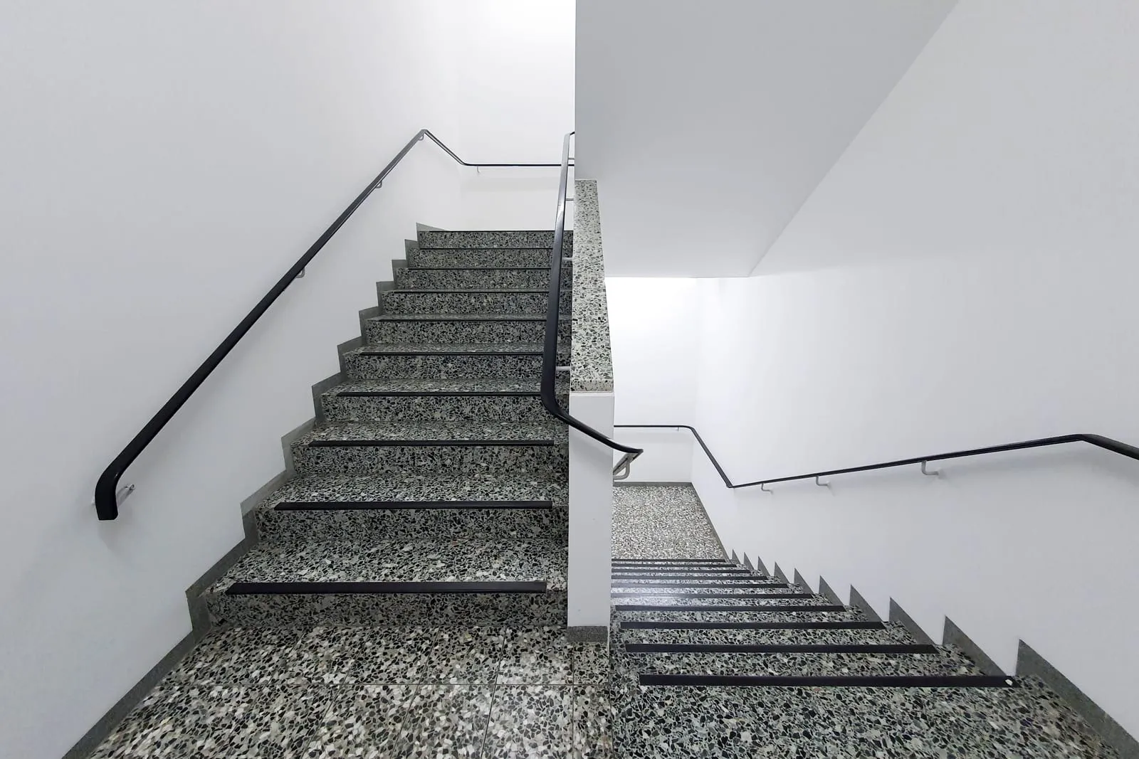 Sauberes Treppenhaus mit Terrazzostufen, weißen Wänden und schwarzem Handlauf
