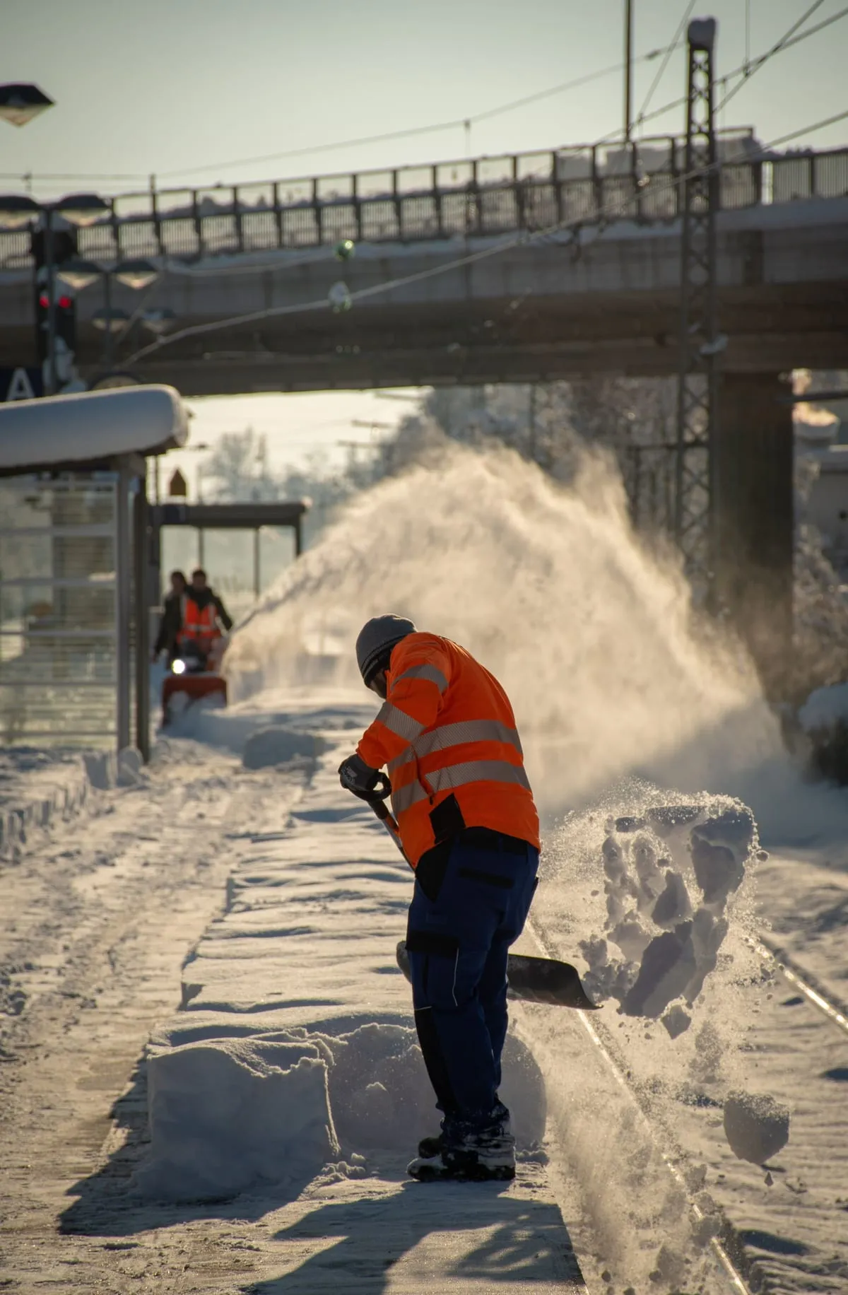 Arbeiter in Warnkleidung schaufelt Schnee auf einem Bahnsteig, von hinten gesehen